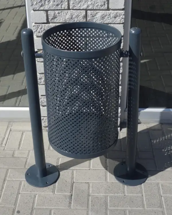 Dual leg swing steel bin — a perforated steel bin body slung between two powder-coated posts on bolt-down base plates, on block paving outside a shopfront