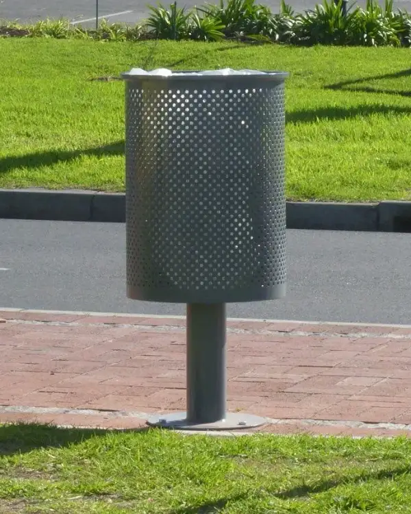 Perforated steel pedestal bin on a bolted baseplate, standing on brick paving beside a kerb and grass verge