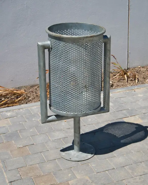 Perforated swing steel bin — a galvanised perforated bin body pivoting in a yoke on a single post with a round bolt-down base plate, on block paving