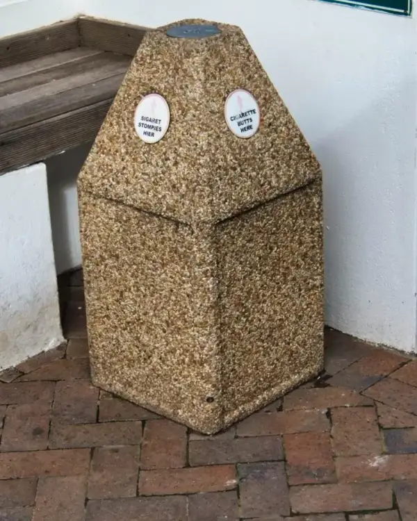Pyramid ash concrete bin in exposed aggregate with a steel stubbing plate at the apex and bilingual cigarette-butt decals, on brick paving