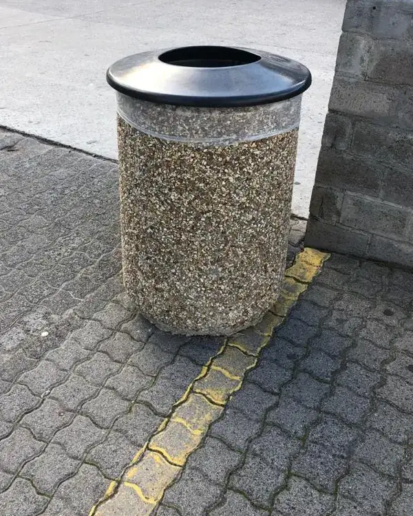 Round 500 concrete bin with a black removable lid with a central aperture, a bag liner folded over the rim beneath it, on forecourt paving