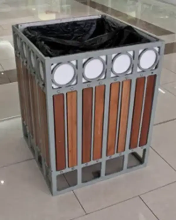 Timber slatted square bin — timber slat infill panels in a grey powder-coated steel frame with a band of ringed discs around the top, open top with a black liner, standing on a tiled floor
