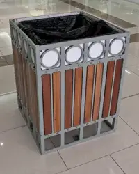 Timber slatted square bin — timber slat infill panels in a grey powder-coated steel frame with a band of ringed discs around the top, open top with a black liner, standing on a tiled floor