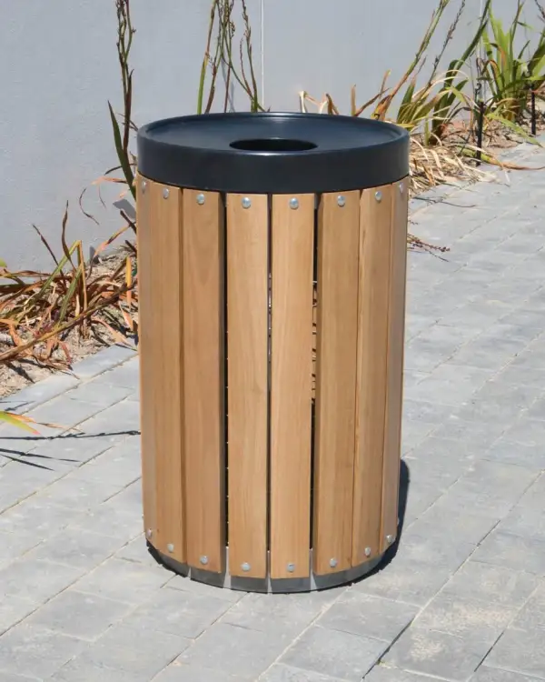 Round timber slatted steel bin — hardwood slats on a galvanised steel frame with galvanised bands and a black moulded lid with a central aperture, standing on block paving beside a planted bed